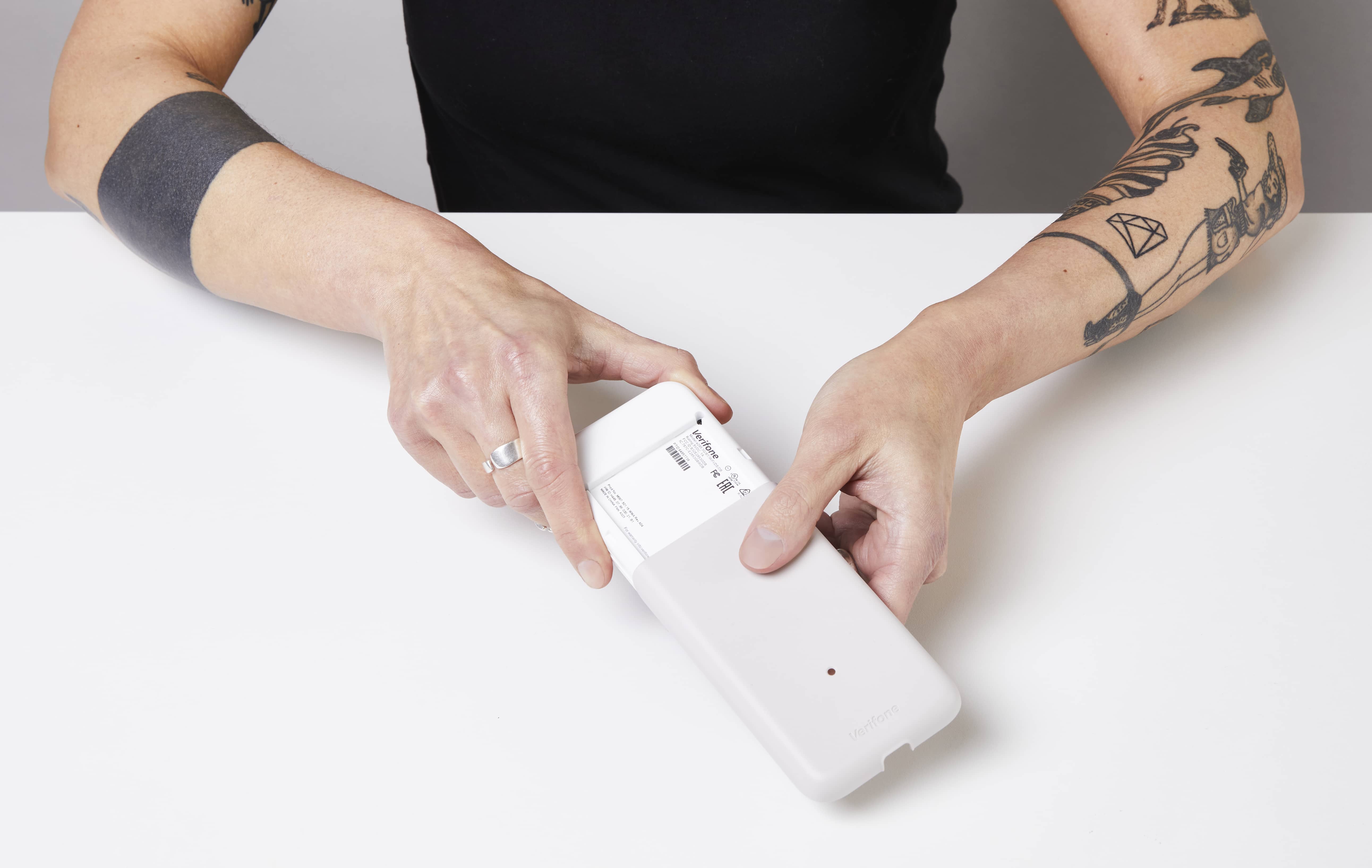 Image montrant deux mains qui tiennent le terminal de paiement e285 face vers le bas. Une main fait glisser le couvercle arrière vers le bas, faisant ainsi apparaître la batterie et l’emplacement de la carte SIM.