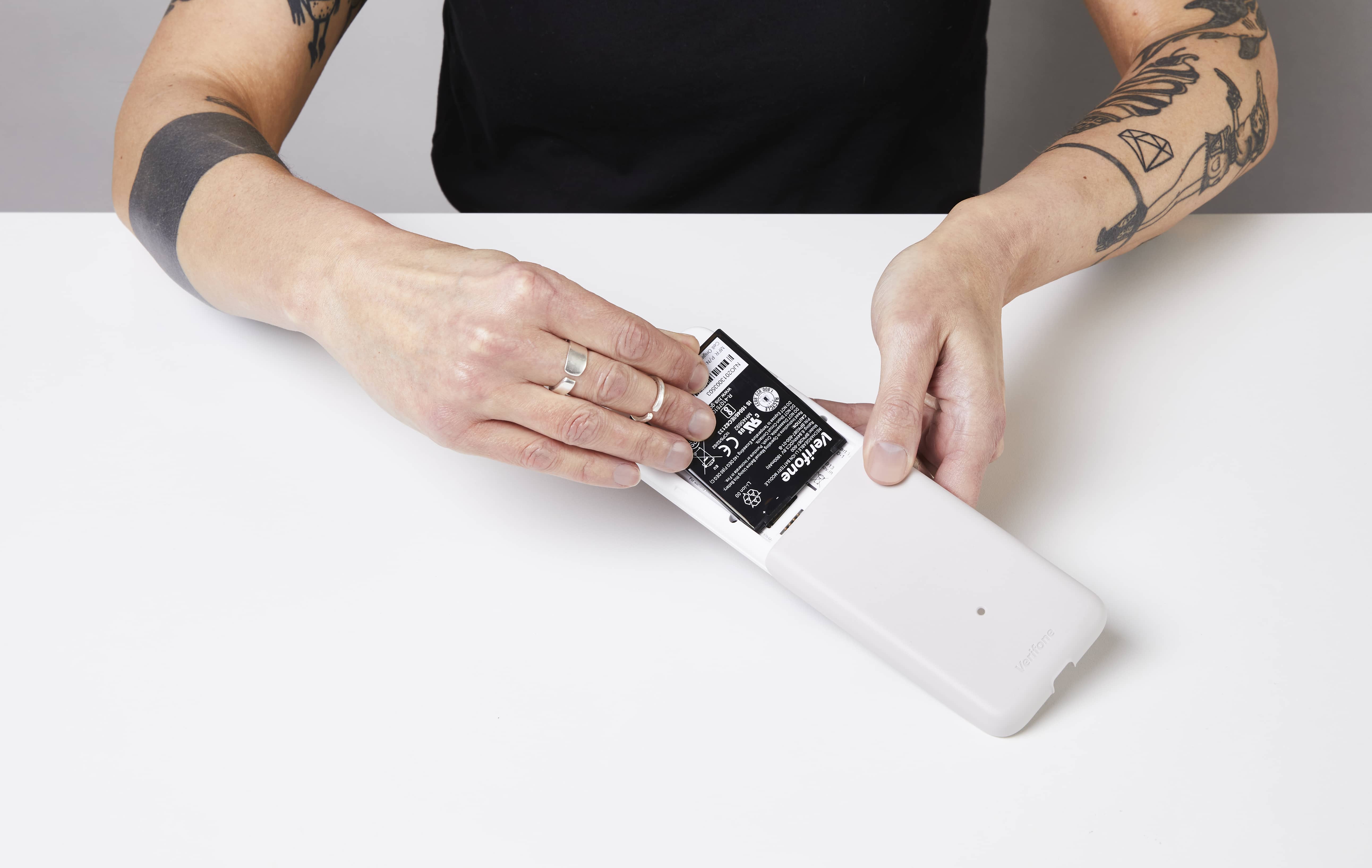 Das Bild zeigt zwei Hände, die das Zahlungsterminal e285 mit der Vorderseite nach unten halten, wobei die hinter Abdeckung abgenommen ist. Eine Hand setzt eine Batterie ein.